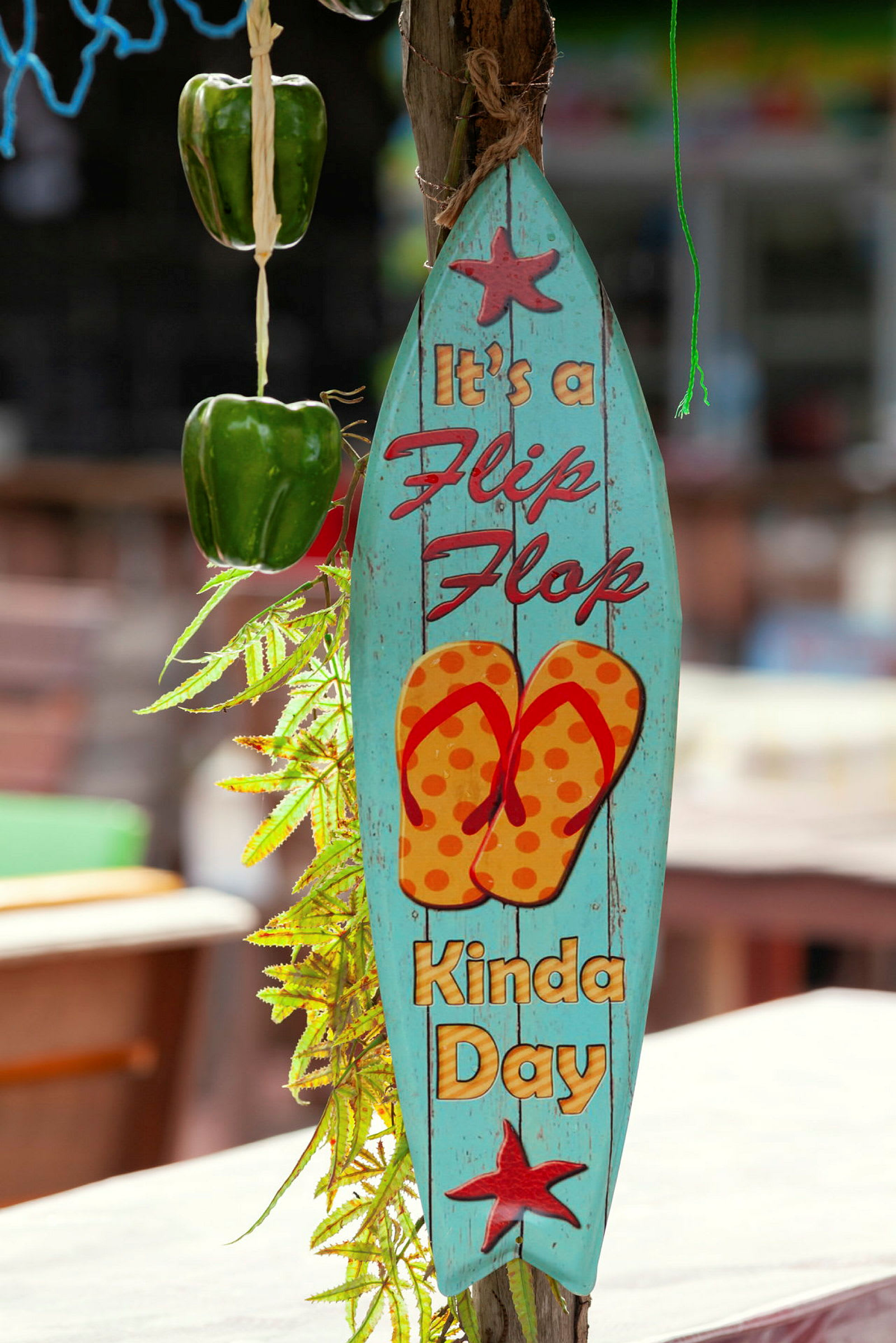 Sininen kyltti, jossa lukee 'It's a flip-flop kinda day', kuva sandaaleista ja koristeltu tähdillä, roikkuu vihreän paprikan ja vihreiden lehtien vieressä. (Tekoälyn luoma teksti)