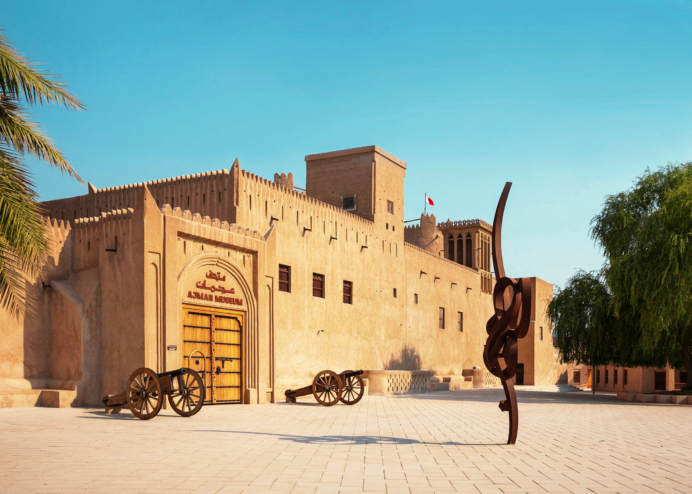 Historiallinen rakennus hiekanvärisillä julkisivuilla, "Ajman Museum" lukee suuren puuoven yläpuolella. Rakennuksen edessä on kaksi tykkiä ja moderni veistos. (Tekoälyn luoma teksti)