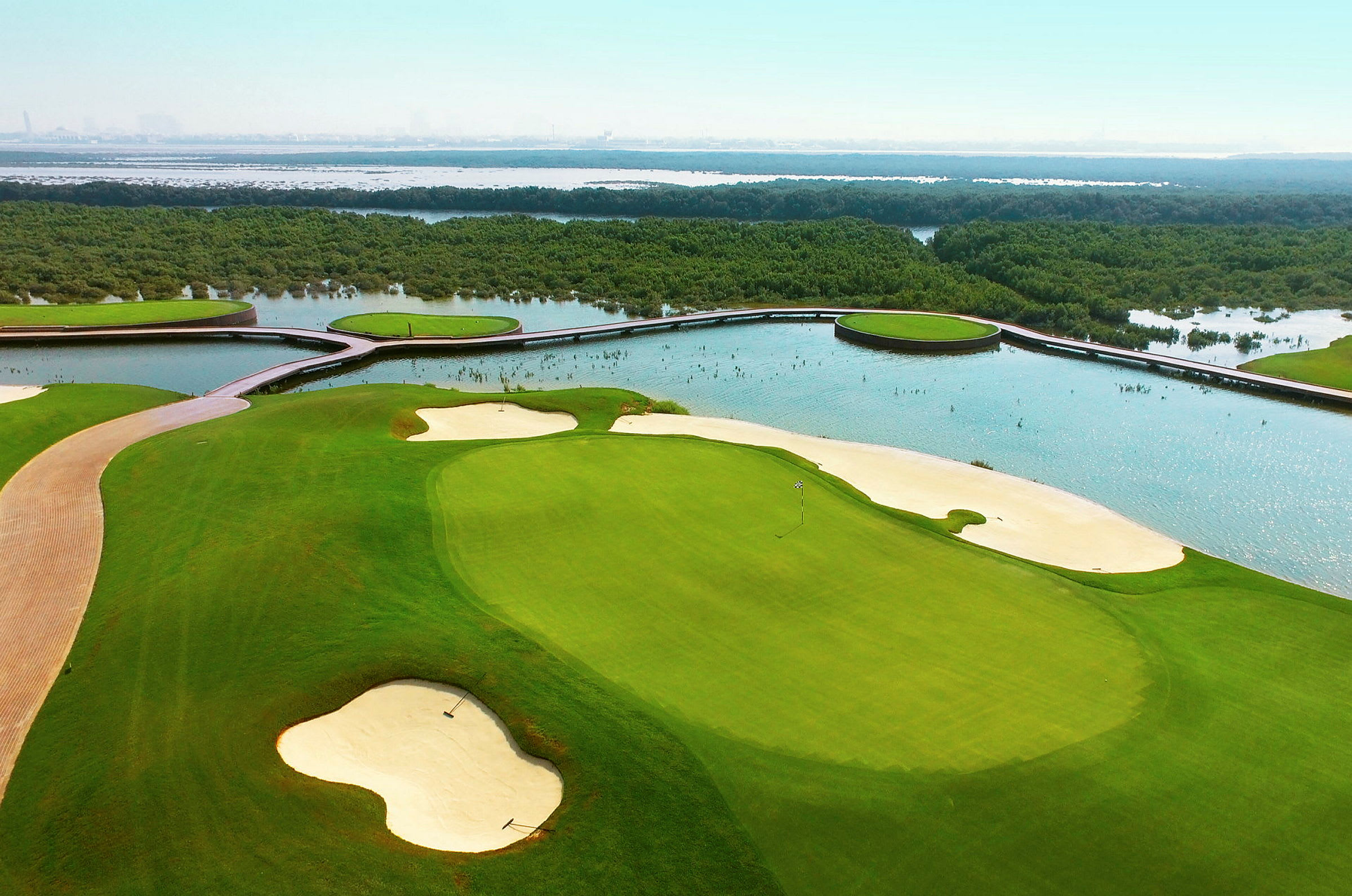 Kuvassa on golfkenttä, jossa on hiekkabunkkereita ja vihreitä väyliä, ympäröitynä vedellä ja kasvillisuudella. (Tekoälyn luoma teksti)