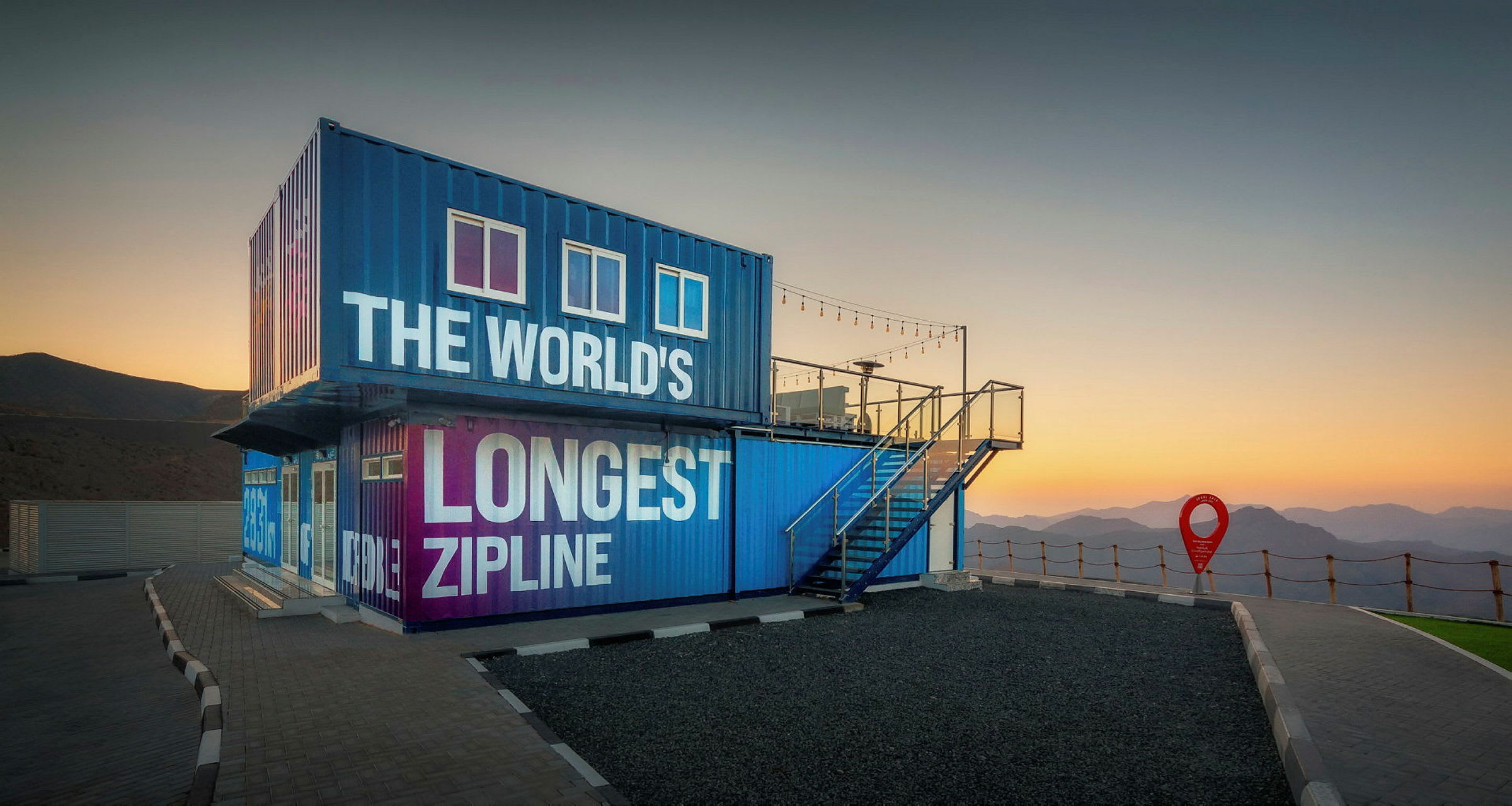 Sininen ja violetti rakennus, jossa lukee 'The World's Longest Zipline', auringonlaskun aikaan, vuorten ympäröimänä. (Tekoälyn luoma teksti)