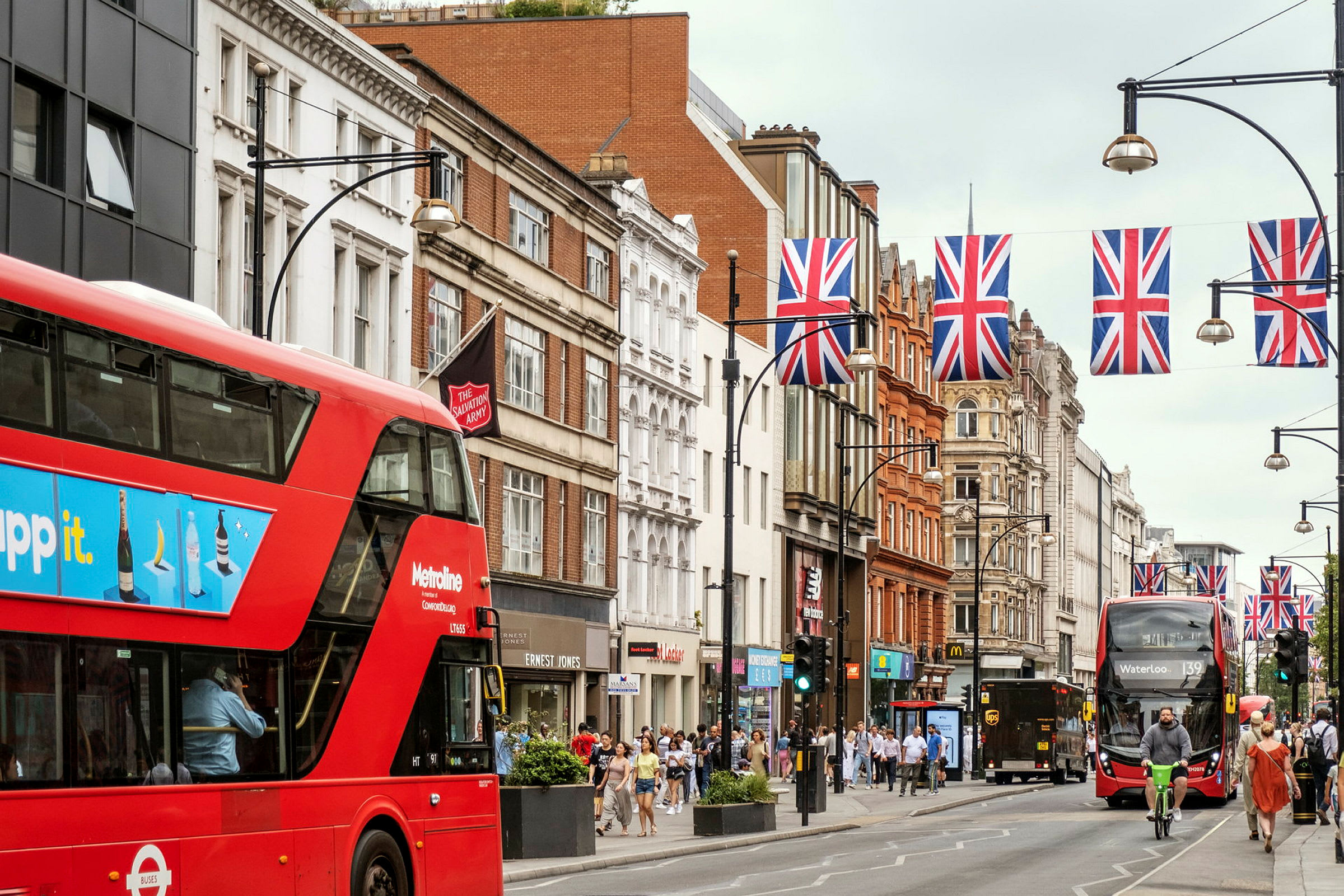Katu punaisilla busseilla, rakennuksia ja yläpuolella roikkuvia Britannian lippuja. (Tekoälyn luoma teksti)