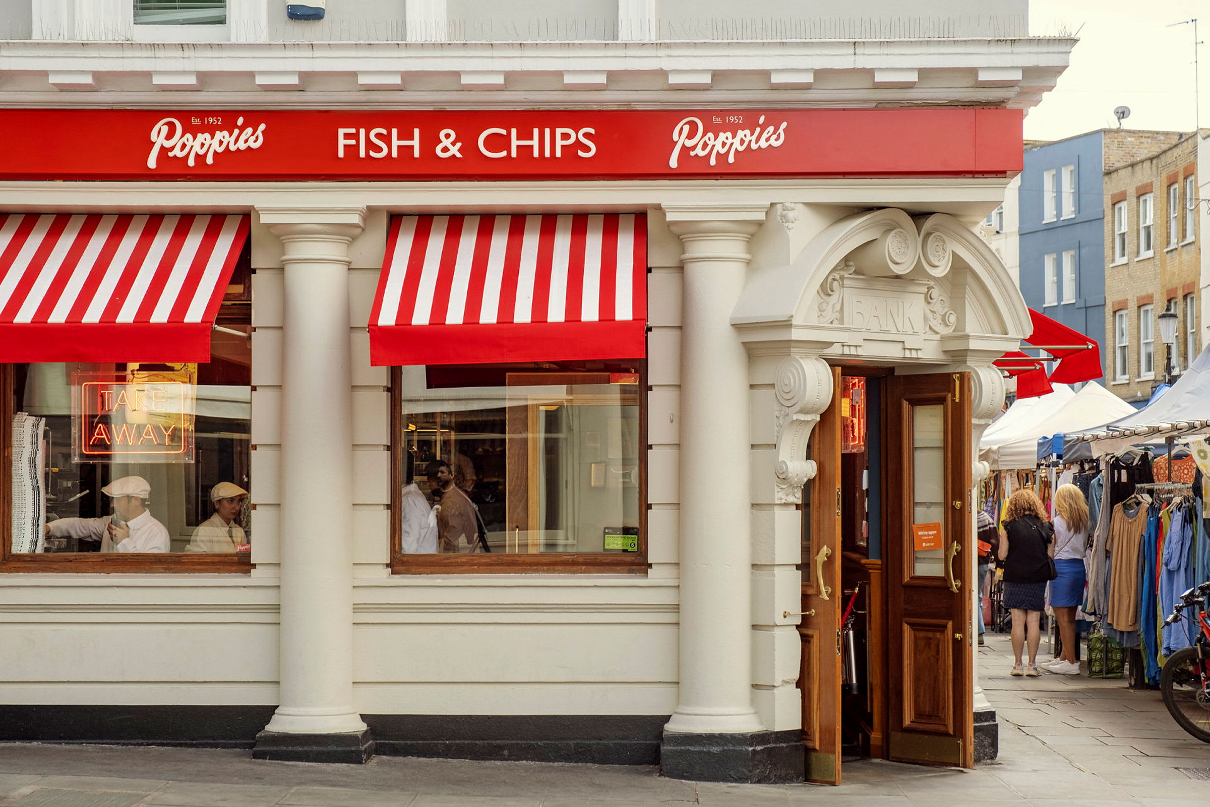 Ravintolan julkisivu, jossa on punaiset ja valkoiset markiisit, 'Fish & Chips' -kyltti ja avoimet ovet. (Tekoälyn luoma teksti)