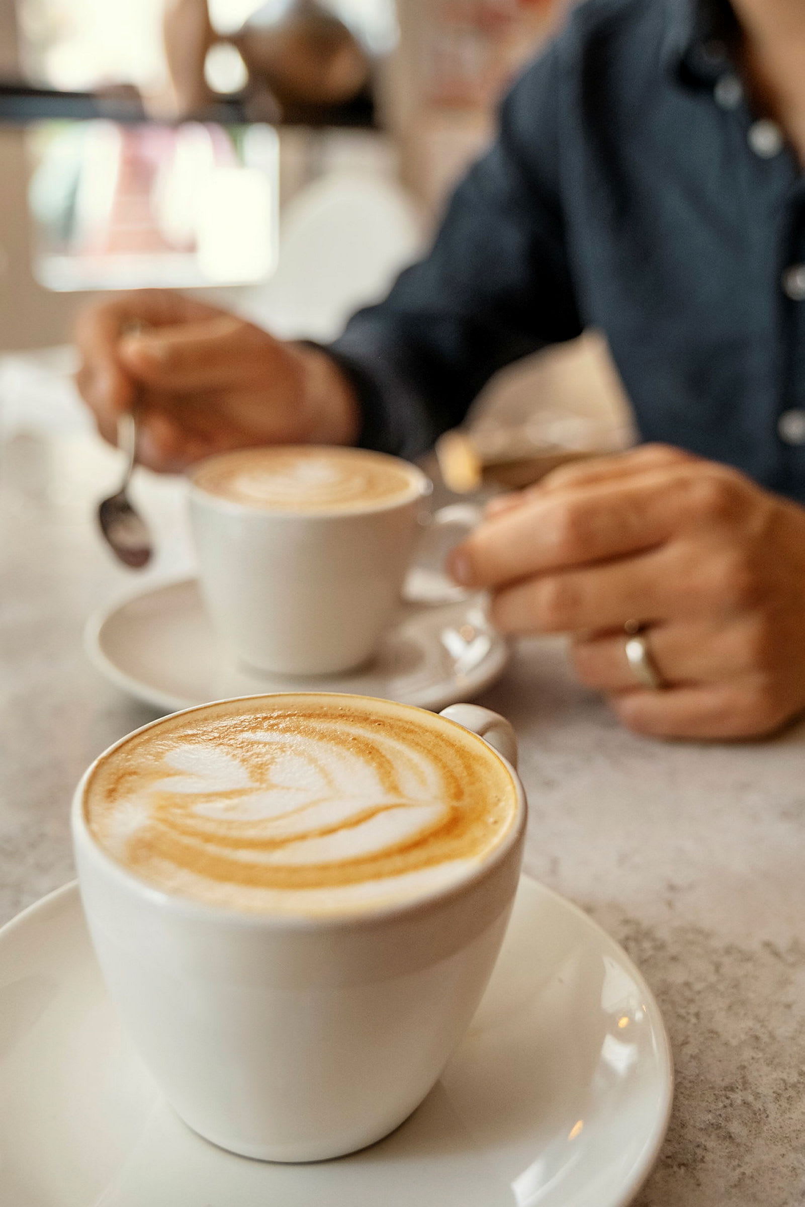 Kaksi cappuccinoa valkoisissa kupeissa pöydällä, käsi pitää lusikkaa. (Tekoälyn luoma teksti)