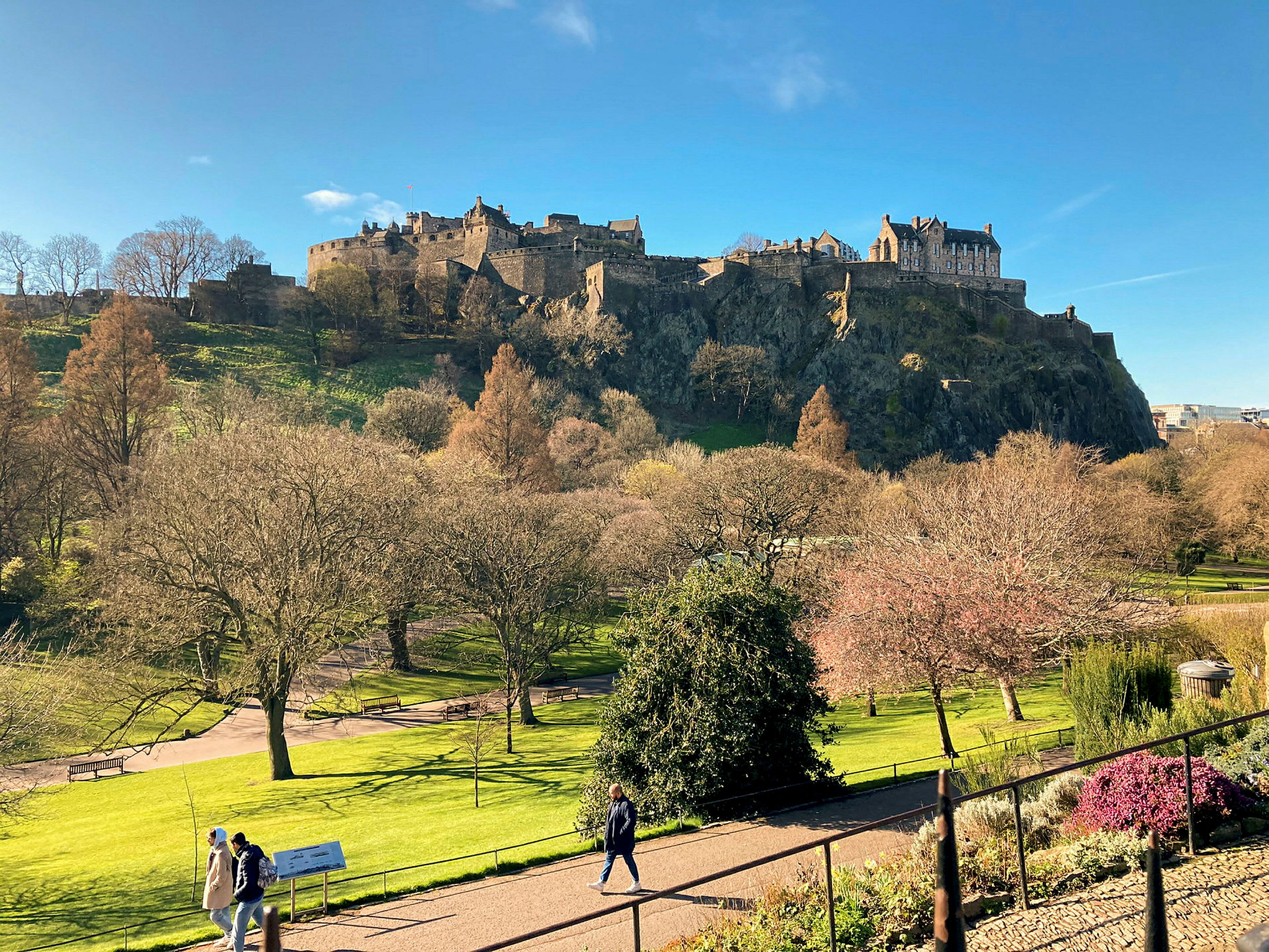 Näkymä Edinburghin linnaan aurinkoisena päivänä, jota ympäröi puita ja vehreää. (Tekoälyn luoma teksti)
