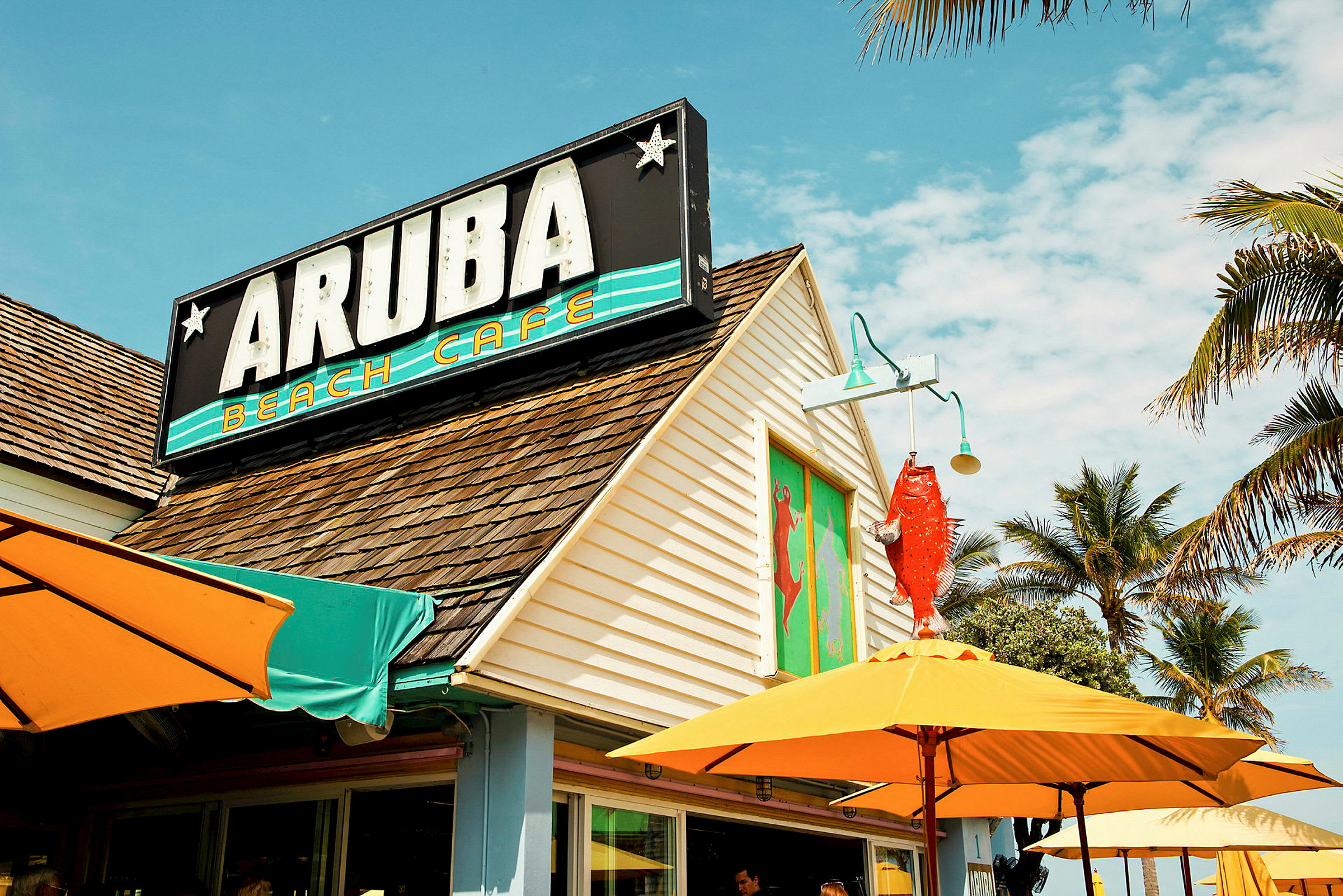 Rakennus, jossa on kyltti "Aruba Beach Cafe" ja ruskeat kattotiilet. Värikkäitä aurinkovarjoja ja palmuja kahvilan ympärillä. (Tekoälyn luoma teksti)