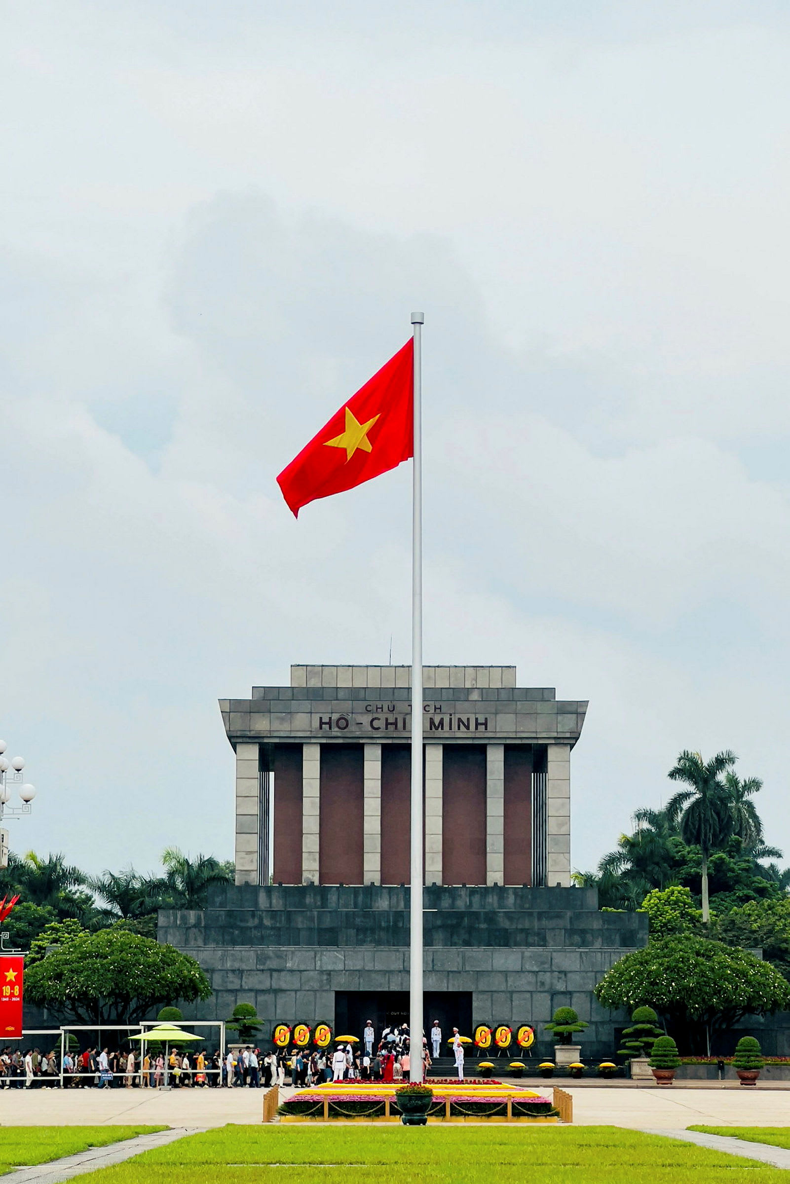 Suuri mausoleumi, jonka julkisivussa on teksti 'Ho Chi Minh', edessä vietnamilainen lippu pystytettynä salkoon. Ihmiset kokoontuvat rakennuksen ympärille, jota ympäröivät puut ja nurmikot. (Tekoälyn luoma teksti)
