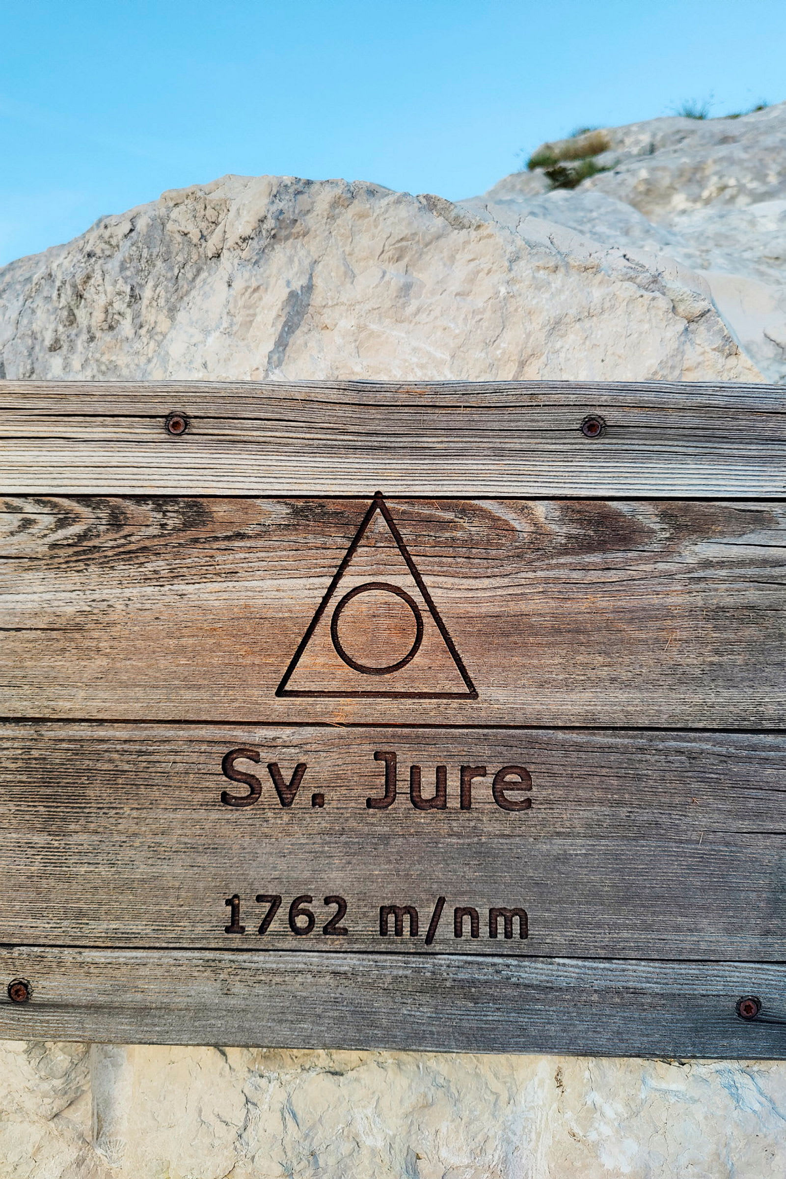 Puukyltti, jossa teksti 'Sv. Jure 1762 m/nm', taustalla kallioita ja sininen taivas. (Tekoälyn luoma teksti)