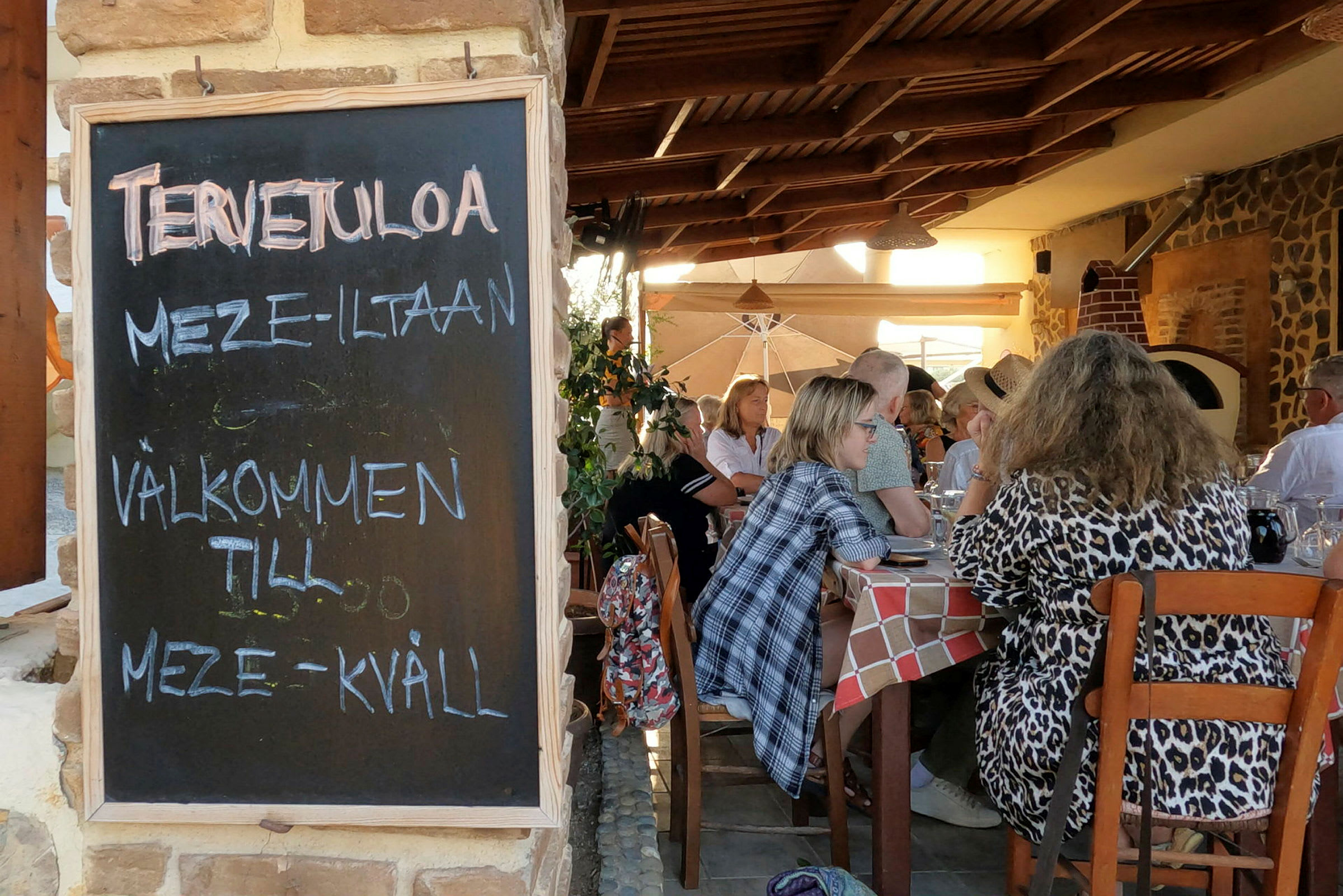 Musta taulu, jossa lukee "Tervetuloa meze-iltaan", sijaitsee ravintolan sisäänkäynnillä. Taustalla näkyy asiakkaita istumassa pöydissä. (Tekoälyn luoma teksti)