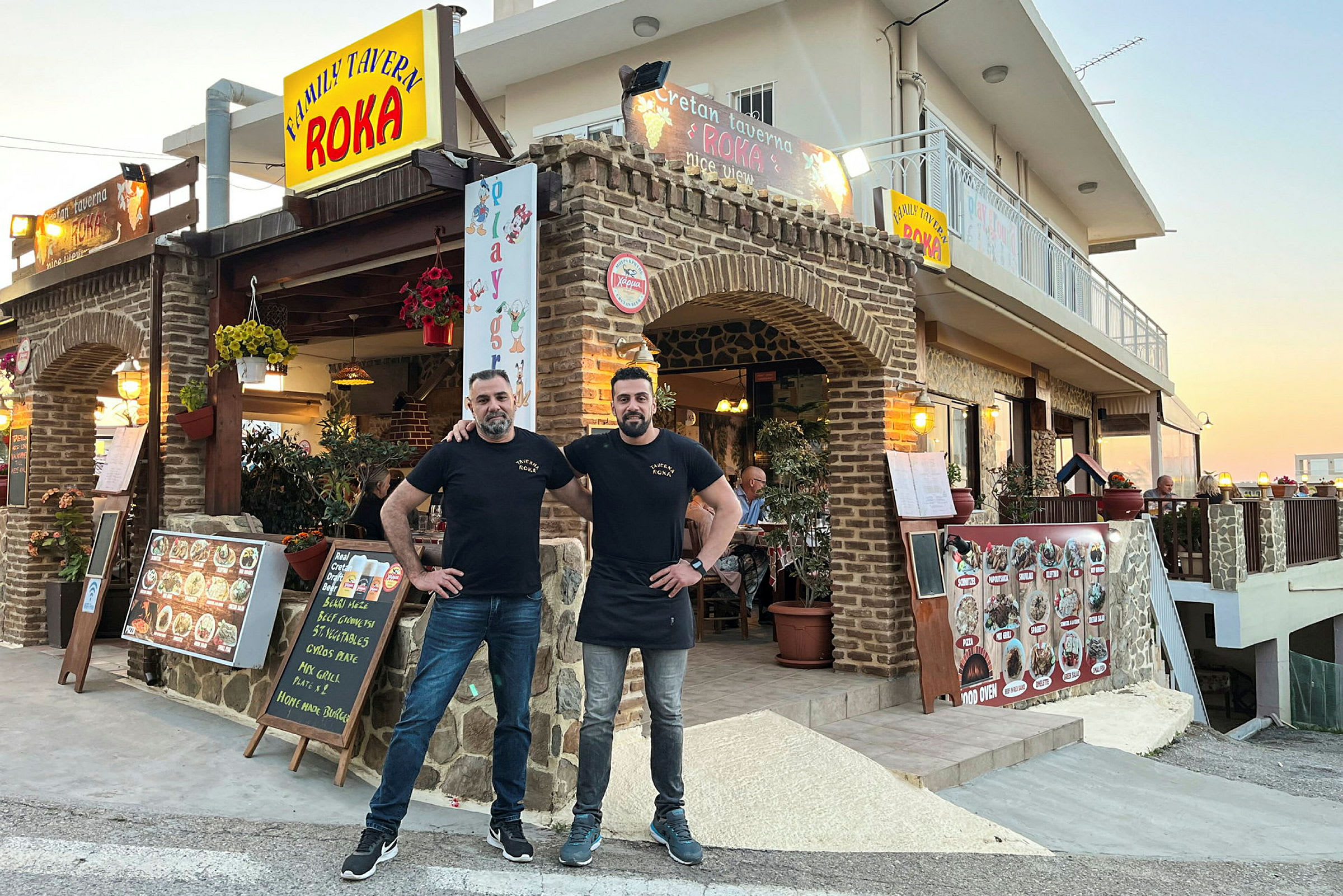 Taverna, joka on rakennettu kivestä, sisältää kylttejä, menuja ja koristeita. Kaksi henkilöä poseeraa edessä. (Tekoälyn luoma teksti)