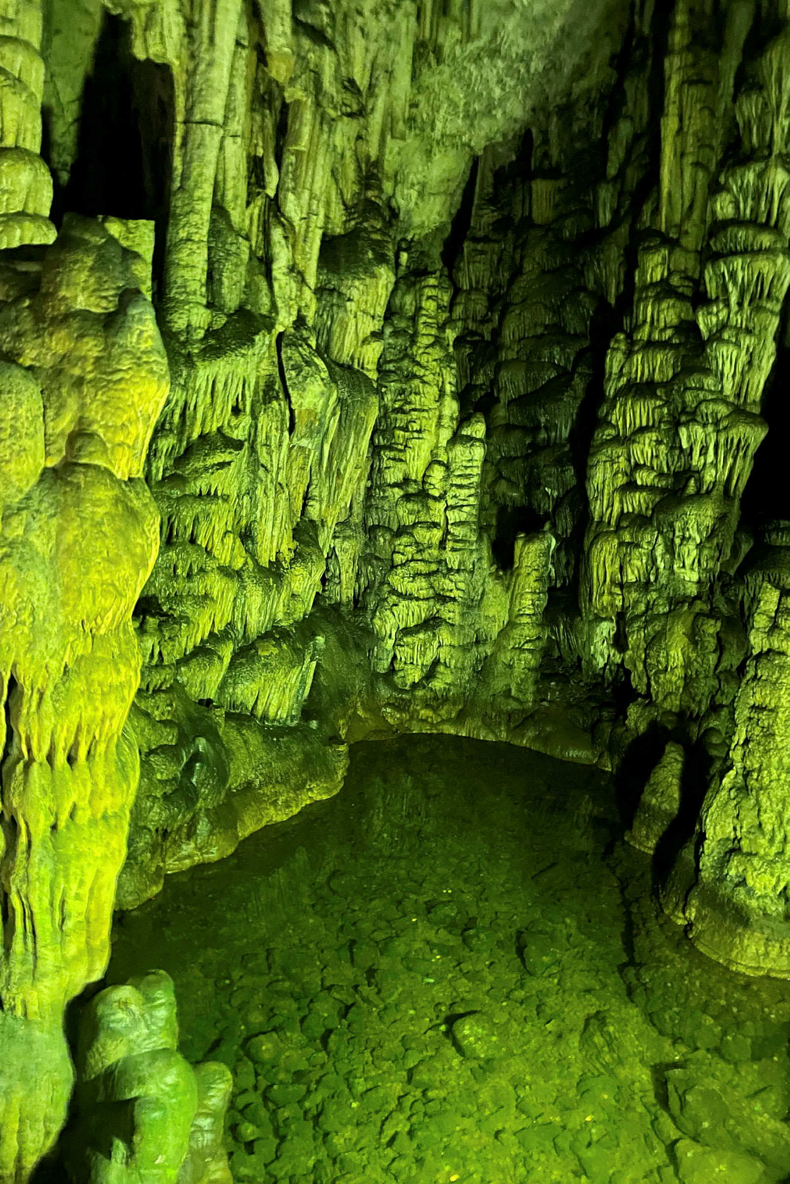 Luola stalaktiittien ja stalagmiittien kanssa, vihreä vesi ja valaistuja kivenmuodostelmia. (Tekoälyn luoma teksti)
