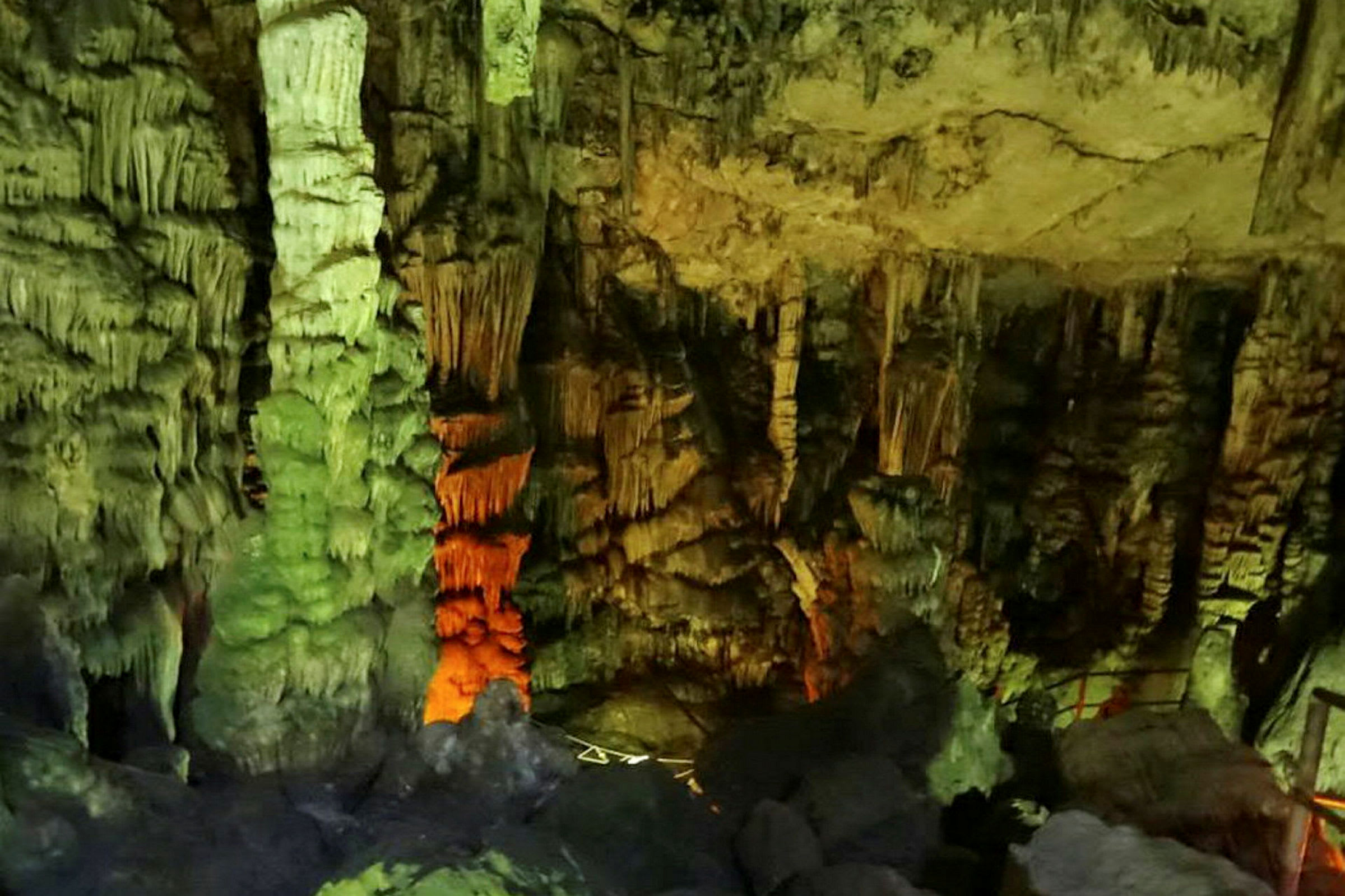 Kuva luolasta, jossa on stalaktiitteja ja stalagmiitteja, valaistu eri väreillä. (Tekoälyn luoma teksti)
