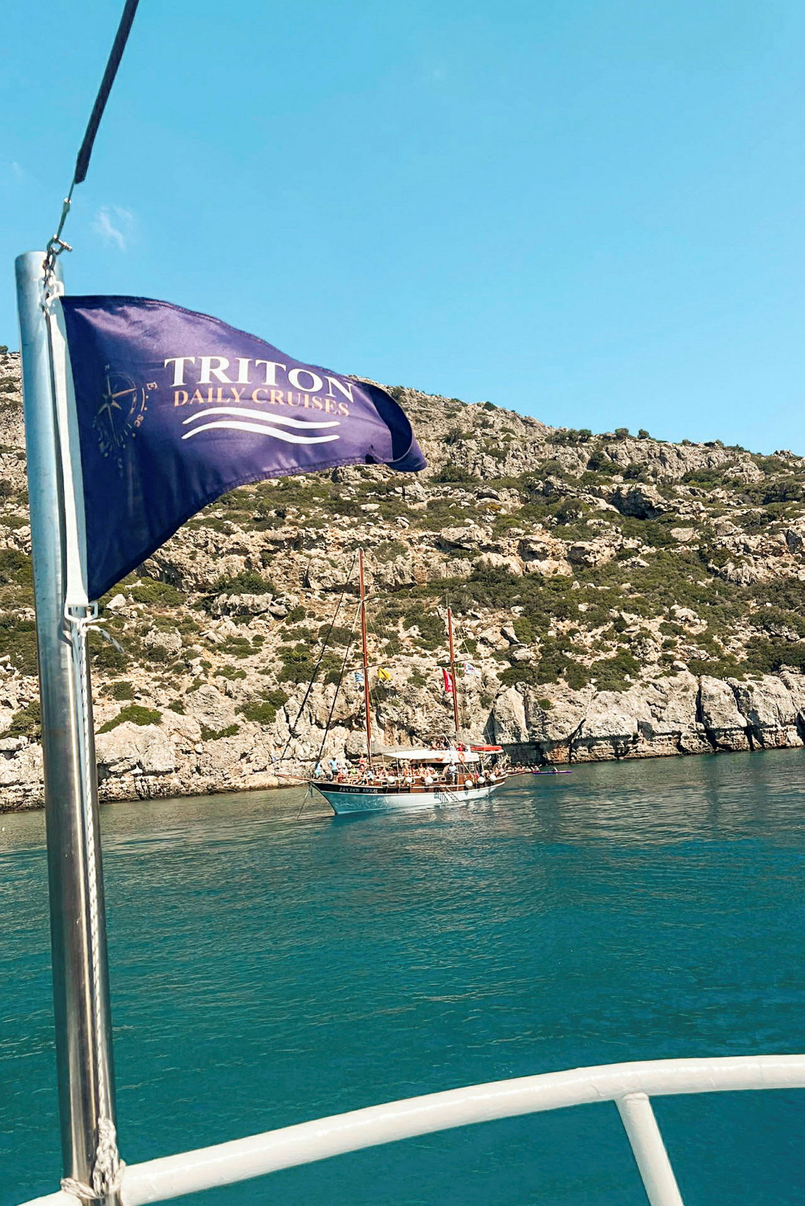 Purjealus on ankkuroitunut kallion viereen, ja etualalla liehuu lippu, jossa lukee "Triton Daily Cruises". (Tekoälyn luoma teksti)