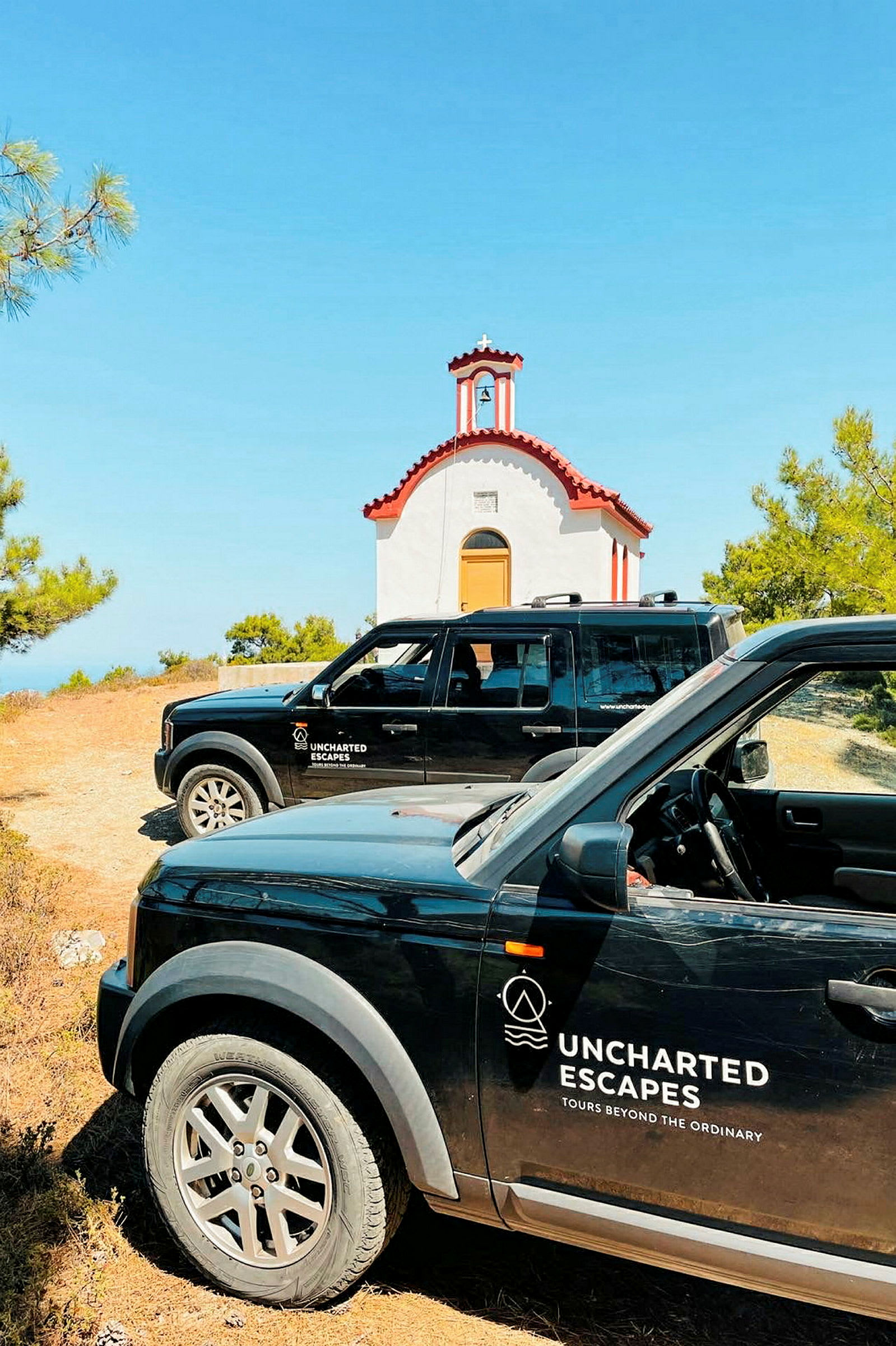Kaksi mustaa autoa, joissa lukee "Uncharted Escapes", on pysäköity pienen, valkoisen kirkon eteen, jossa on punainen katto. Kirkko on vehreyden ympäröimä ja taivas on kirkkaansininen. (Tekoälyn luoma teksti)