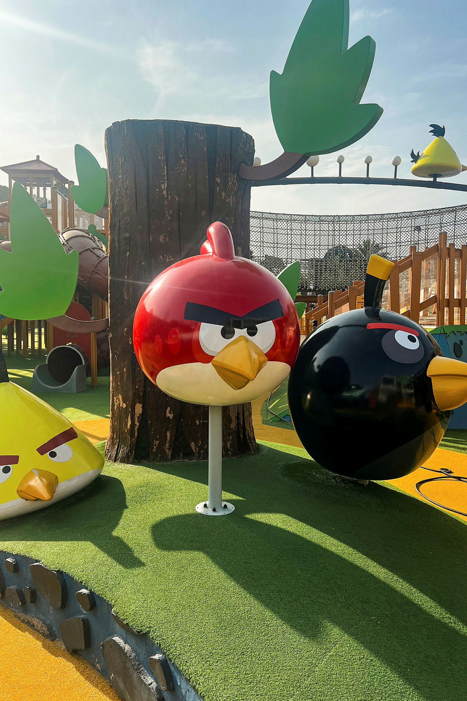 Leikkipaikka Angry Birds -hahmoilla, mukaan lukien punainen lintu, musta lintu ja keltainen kolmionmuotoinen lintu, joissa on puita ja muita leikkirakenteita ympärillä. (Tekoälyn luoma teksti)