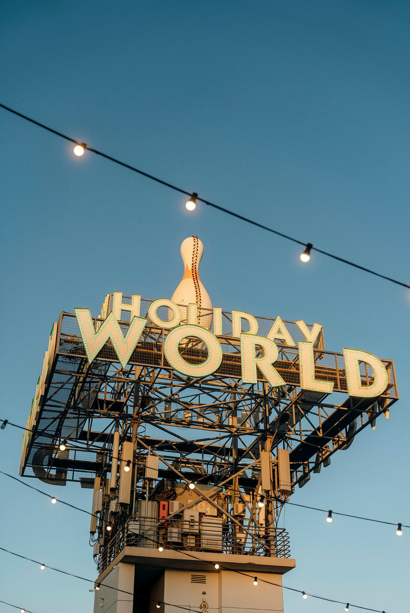 Kyltti, jossa teksti 'Holiday World' ja suuri koristeltu keilapallo yläosassa, valosarjoja kirkasta sinistä taivasta vasten. (Tekoälyn luoma teksti)