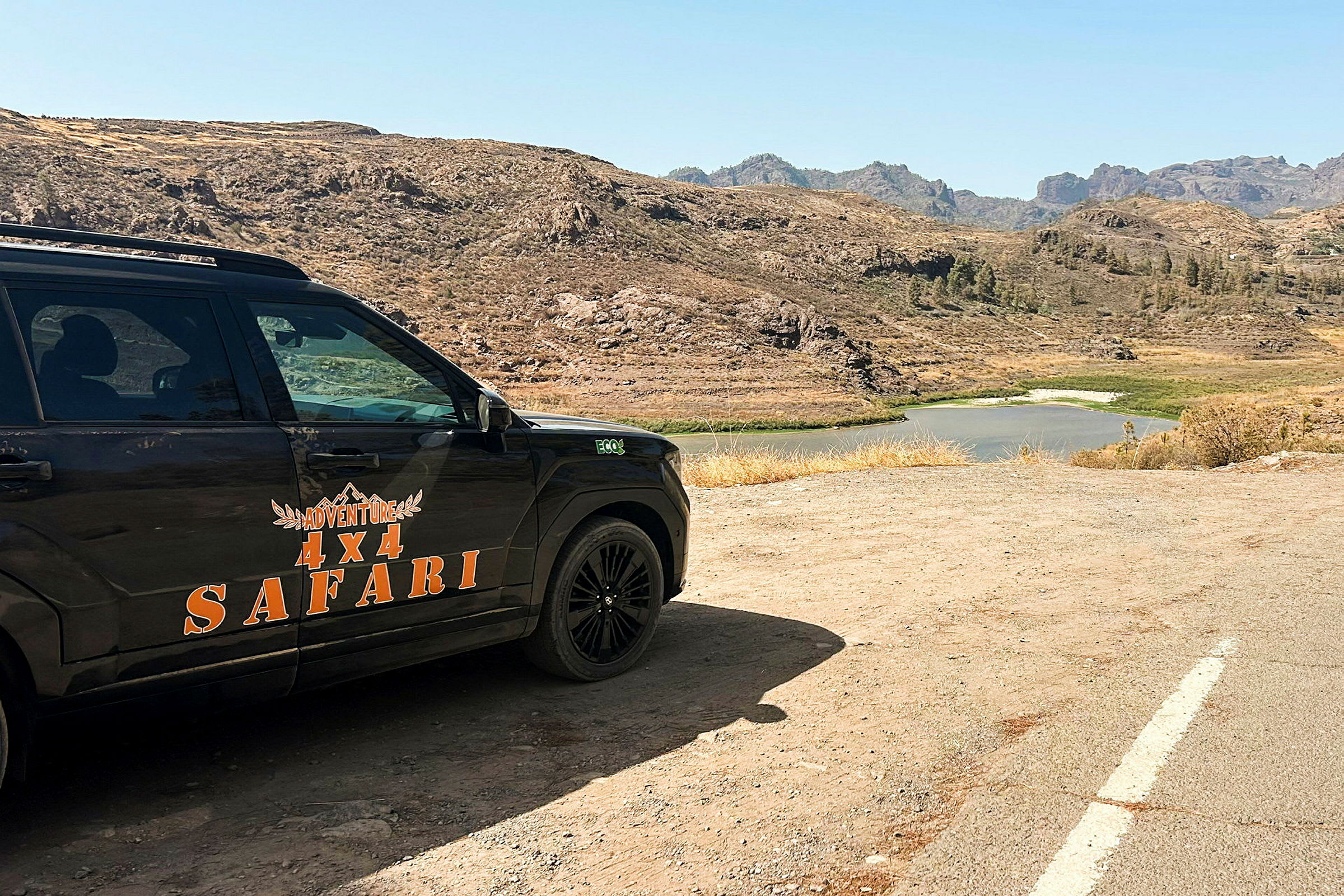 Neljän pyörän vetoauto tekstillä 'Adventure 4x4 Safari', pysäköity autiomaa-maisemaan, jossa taustalla näkyy järvi. (Tekoälyn luoma teksti)