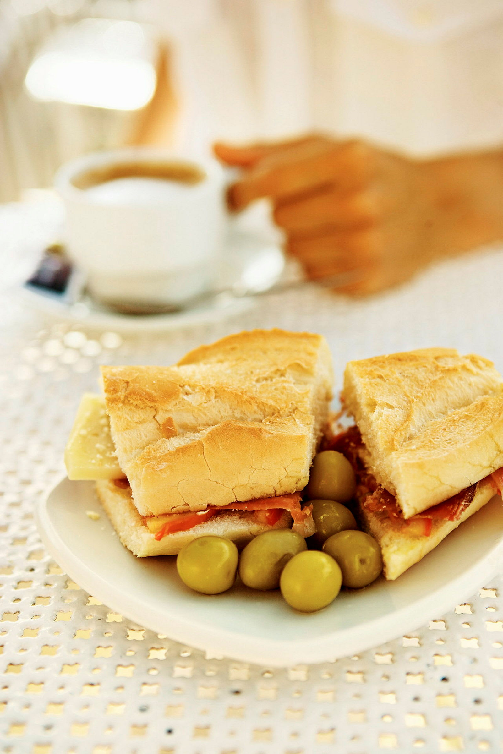 Lautasella on baguette, jossa on juustoa ja kinkkua, sekä muutama oliivi. Taustalla näkyy käsi, joka pitää kahvikuppia. (Tekoälyn luoma teksti)