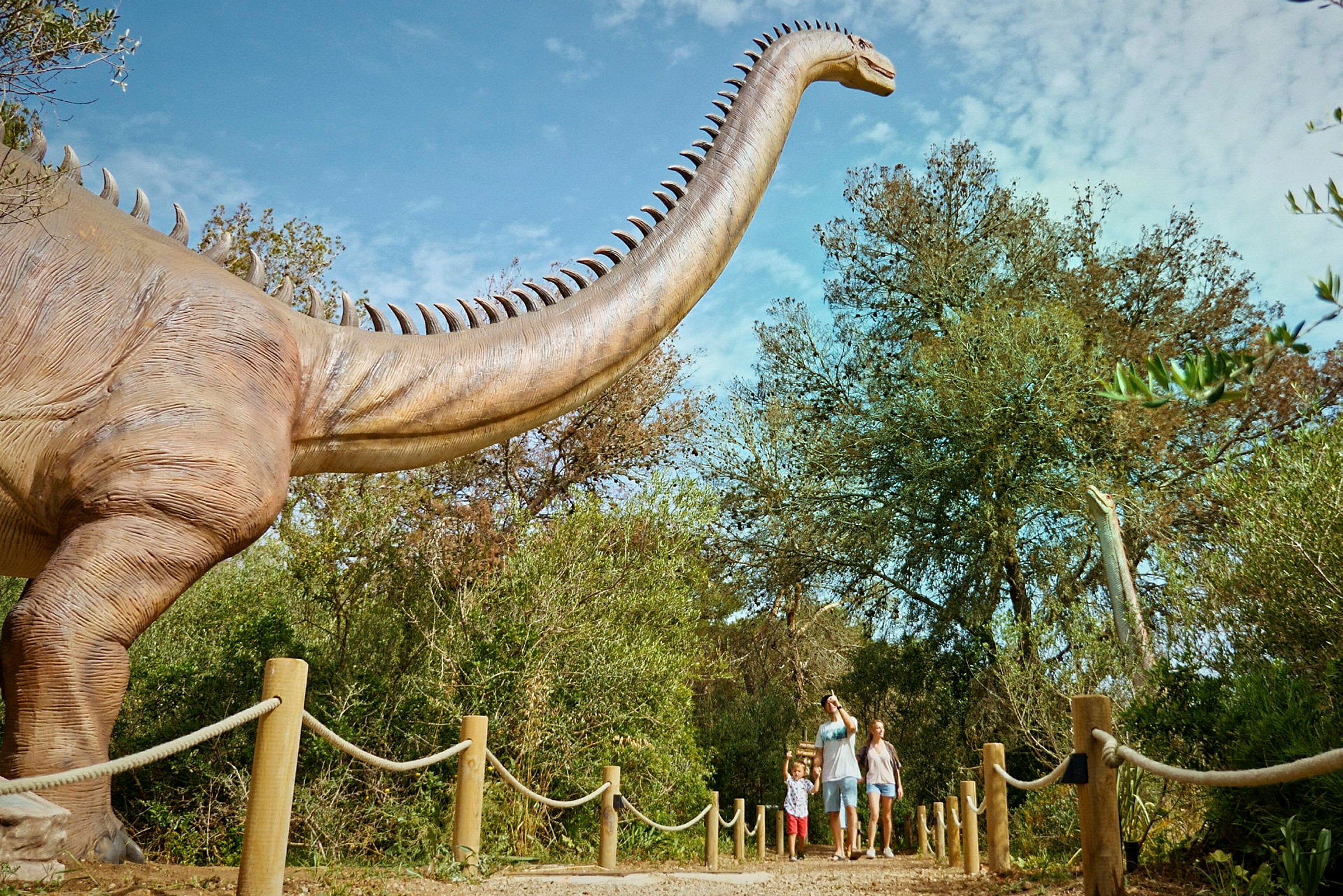 Perhe kävelee polkua pitkin suuren dinosaurusmallin vieressä ulkonäyttelyssä. (Tekoälyn luoma teksti)