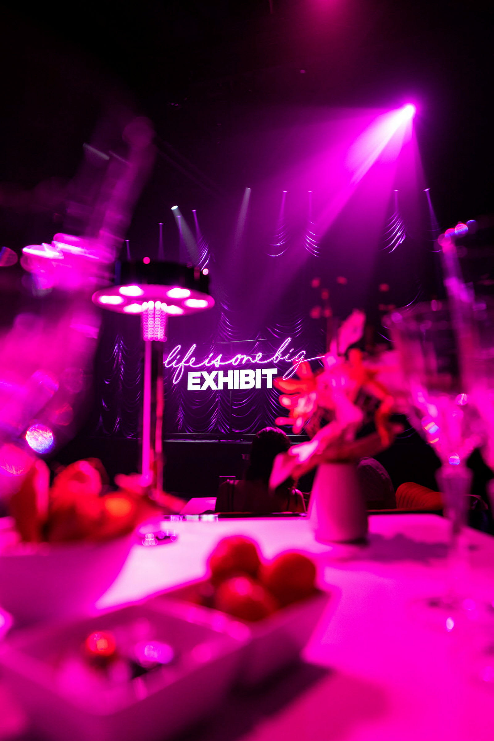 Valaistu näyttämö, jossa on neonvaloilla "life is one big exhibit" -teksti taustalla. Etualalla näkyy pöytä koristeineen ja kulhoineen. (Tekoälyn luoma teksti)