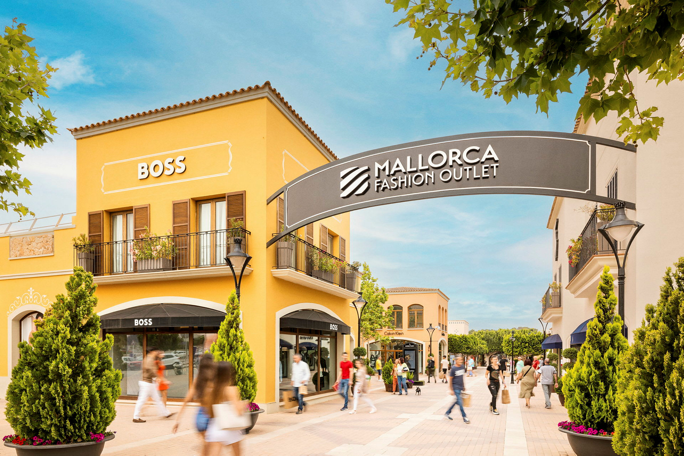 Ostosalue, jossa on keltaisia rakennuksia ja kauppojen kylttejä, kuten BOSS. Sisäänkäynnin yläpuolella näkyy kyltti, jossa lukee "Mallorca Fashion Outlet". (Tekoälyn luoma teksti)