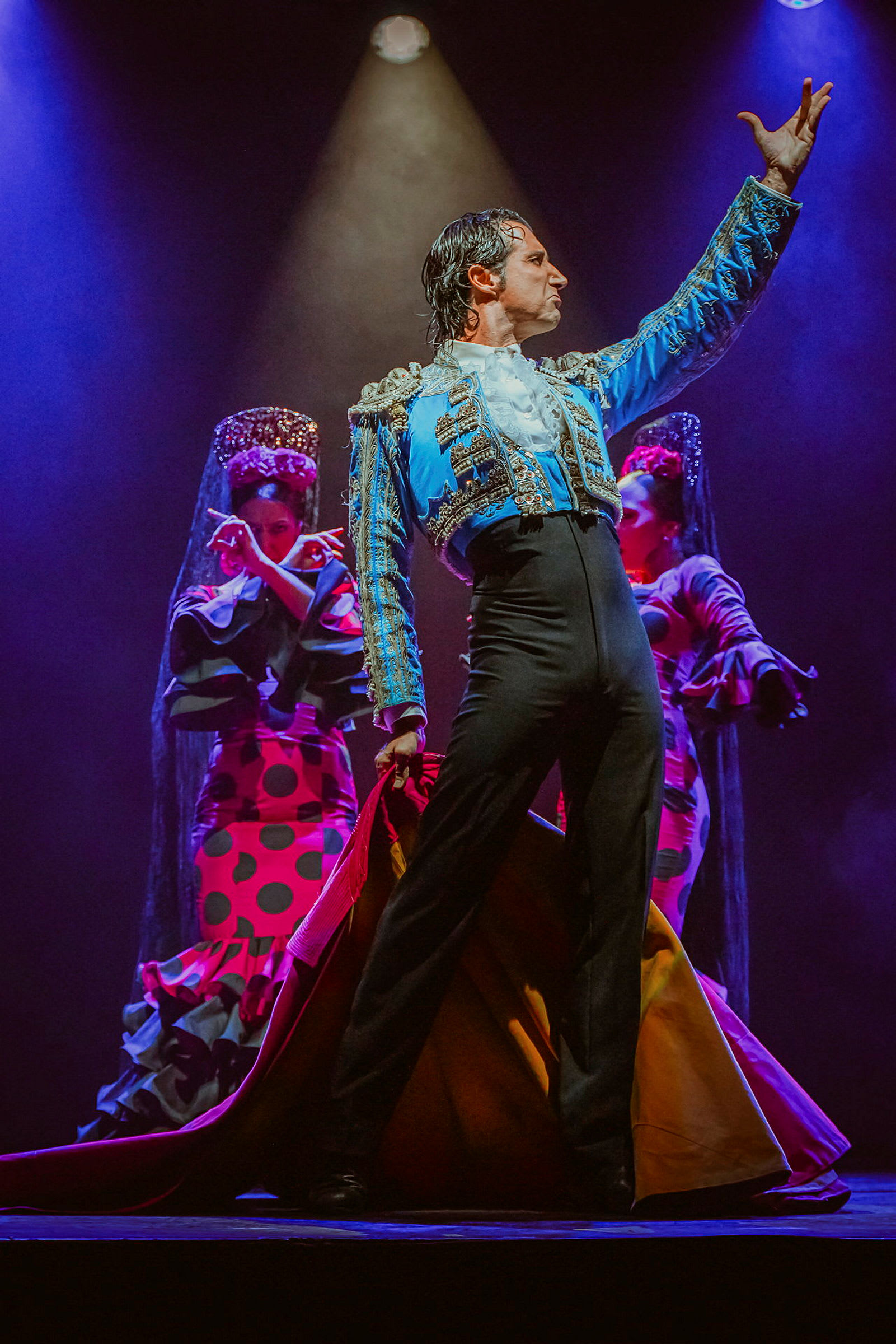 Flamenco-tanssija värikkäässä asussa poseeraa lavalla kahden taustatanssijan kanssa perinteisissä vaatteissa. (Tekoälyn luoma teksti)