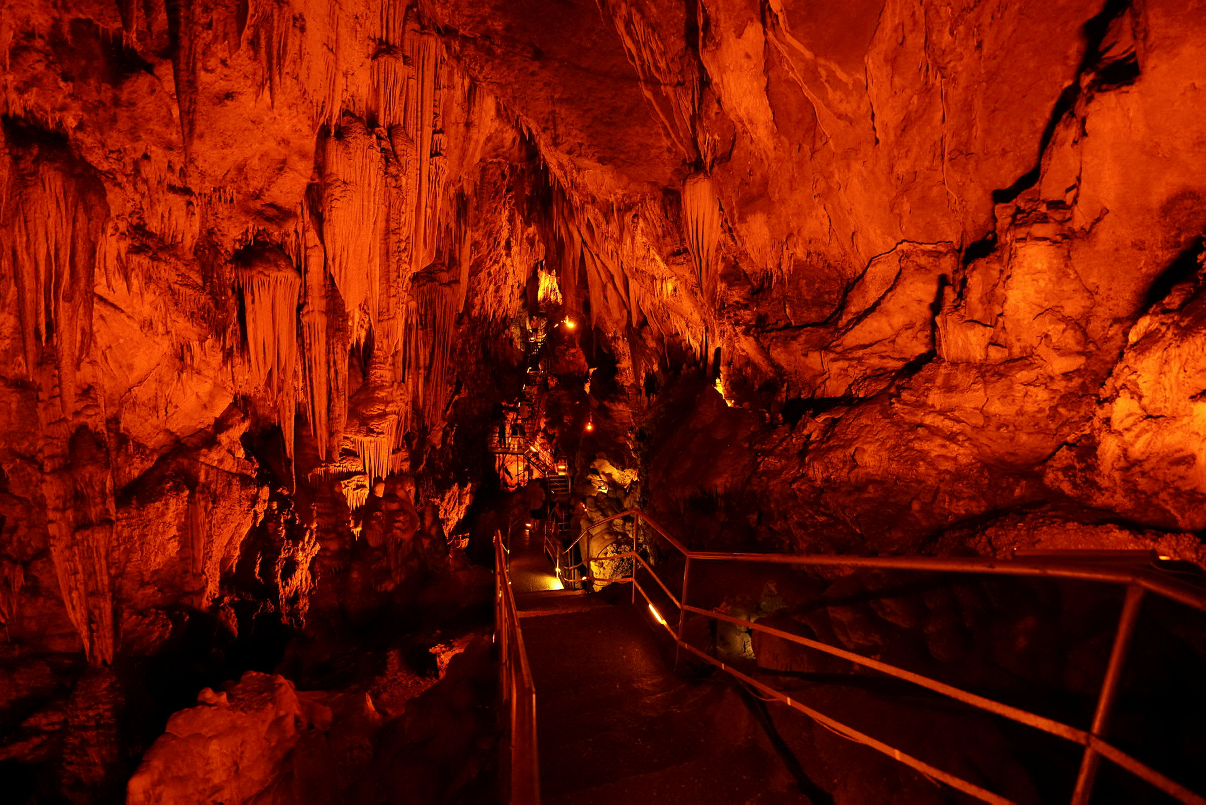 Luola, jossa on stalaktiitteja ja stalagmiitteja, valaistu oranssilla valaistuksella. Kulkutie kaiteineen kulkee luolan läpi. (Tekoälyn luoma teksti)