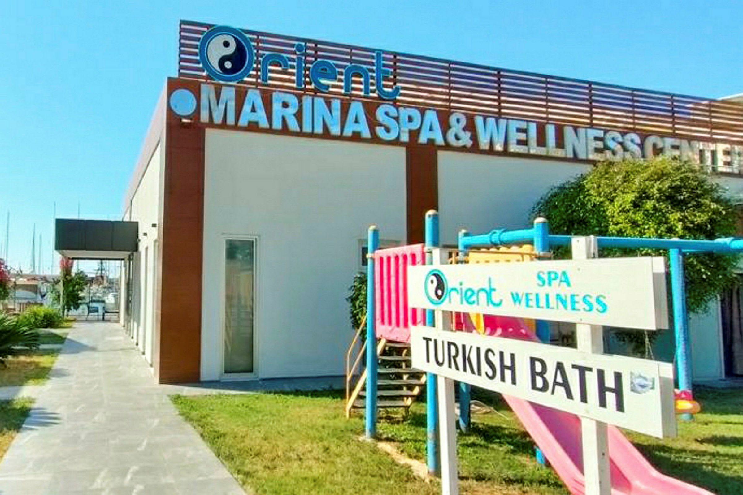 Rakennus, jossa on kyltti 'MARINA SPA & WELLNESS CENTER'. Nurmikolla on myös lasten leikkipaikka ja kyltti tekstillä 'TURKISH BATH'. (Tekoälyn luoma teksti)